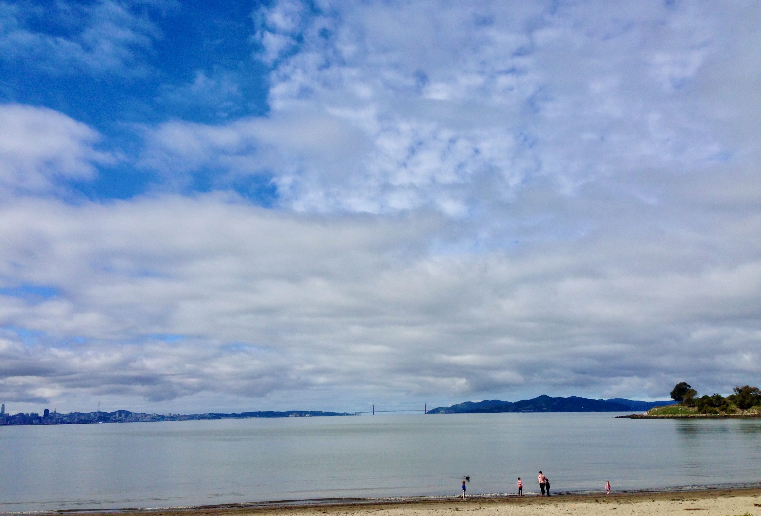 sky with clouds, placid water, some people on the water's edge, city across the water